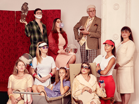 Group portrait of nine people in vintage clothing with an owl, fishbowl, tennis racket, and drinks against a red backdrop.