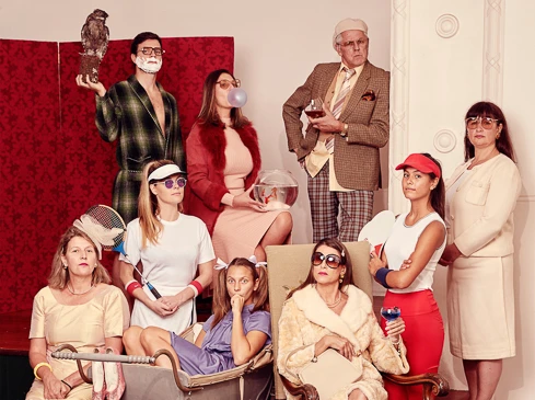 Group portrait of nine people in vintage clothing with an owl, fishbowl, tennis racket, and drinks against a red backdrop.