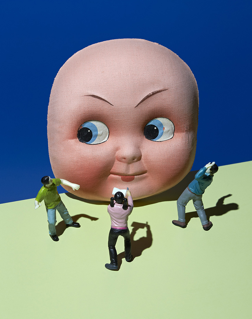 Large doll head looms over miniature people figurines on a split green and blue background in a surreal studio setup.