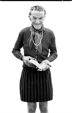 Elderly man in a cardigan and skirt holding a heeled shoe, captured in a monochrome portrait by Chris Budgeon.