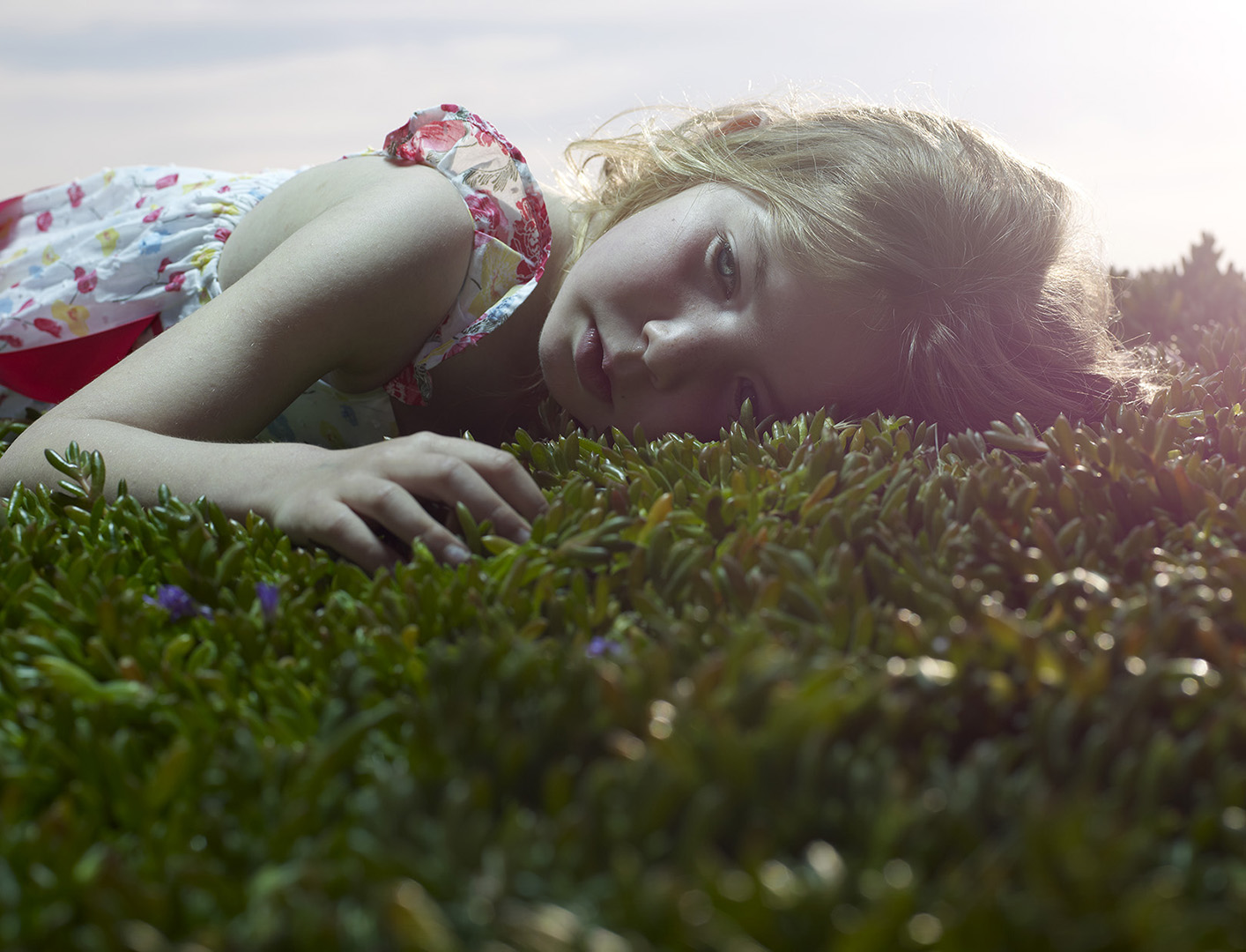 Girl in a floral dress rests in greenery in soft, diffused light, creating a peaceful and dreamy scene, captured in a close-up, eye-level shot.