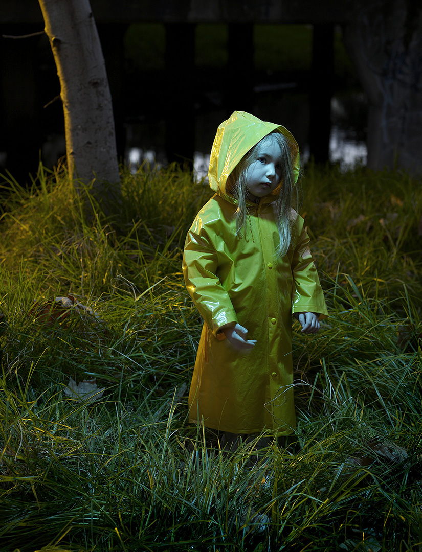 Child in a yellow raincoat stands in tall green grass, illuminated by an unseen light source in a dark outdoor setting. Moody lighting.