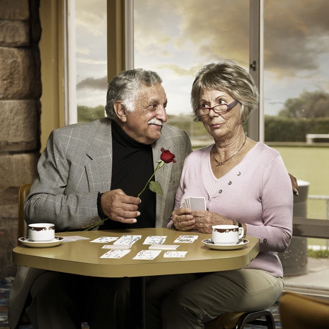 Elderly couple at table with playing cards and coffee, the man holding a red rose, in an indoor setting with natural lighting.