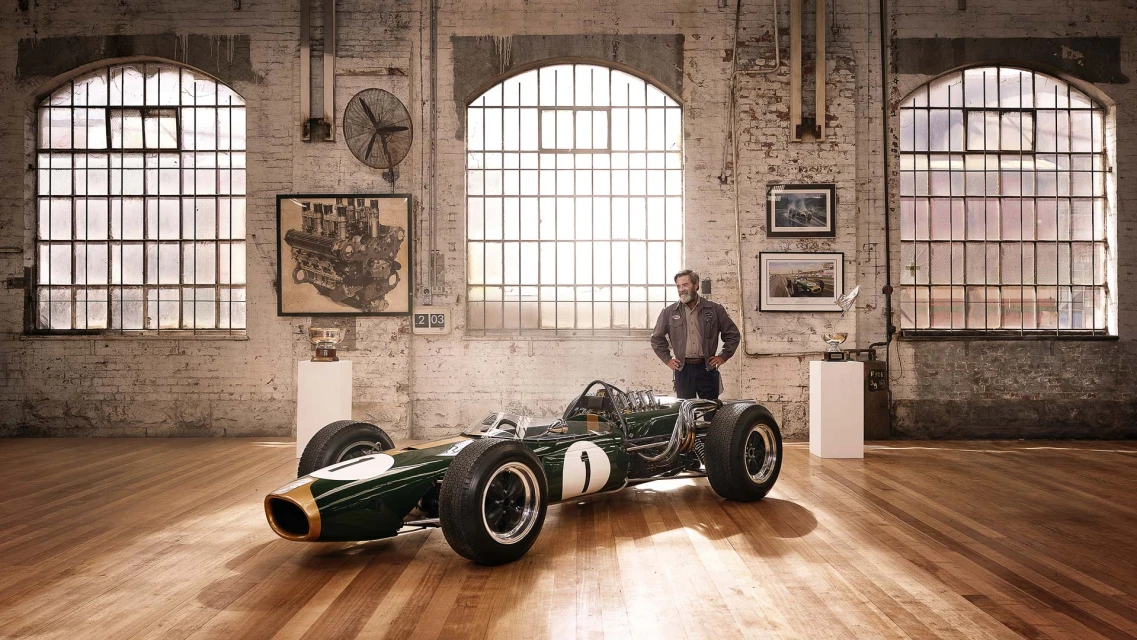 Vintage green and gold race car on wood floor in an industrial-style garage with a man standing nearby. Framed artwork on brick wall.