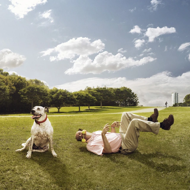 Man lies on grass with tennis balls in his mouth, while a spotted dog sits nearby in a park on a sunny day with trees and blue sky.