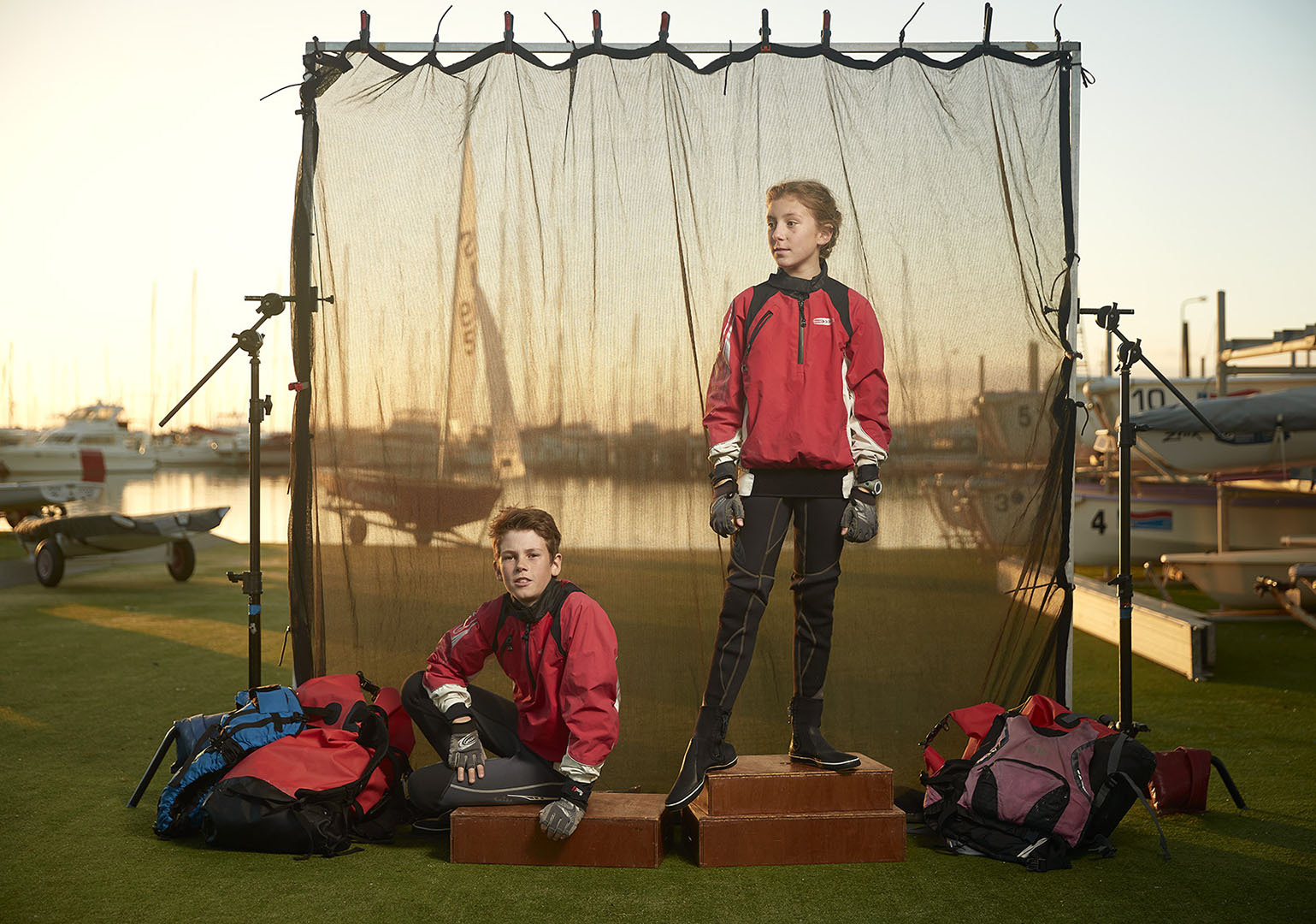 Two young sailors pose outdoors in red jackets and black sailing gear, standing on a low platform against a blurred sailing backdrop.