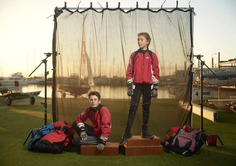 Two young sailors pose outdoors in red jackets and black sailing gear, standing on a low platform against a blurred sailing backdrop.