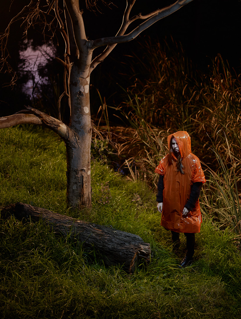 A person in an orange hooded raincoat stands near a fallen log in a grassy field, with a bare tree and tall grasses in the background.