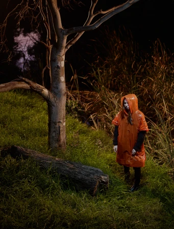 A person in an orange hooded raincoat stands near a fallen log in a grassy field, with a bare tree and tall grasses in the background.