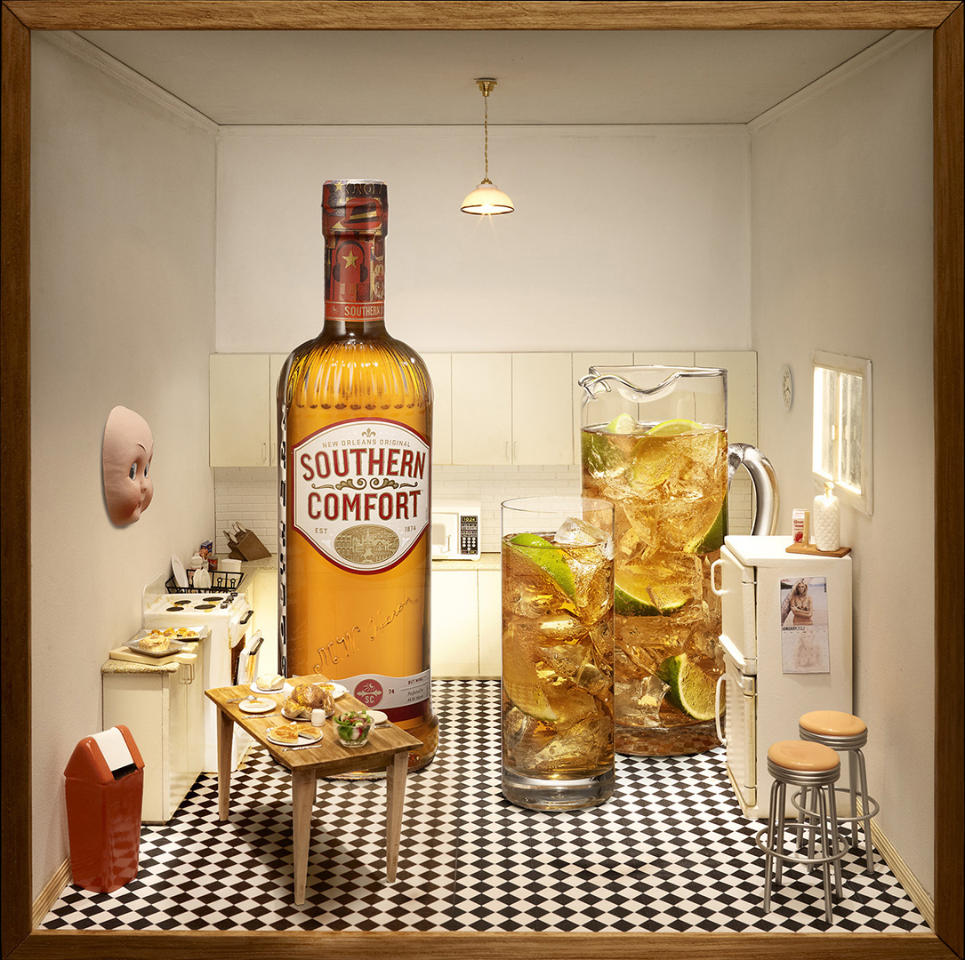 Southern Comfort whiskey bottle is the focal point of a miniature kitchen scene with iced drinks and doll parts on the wall.