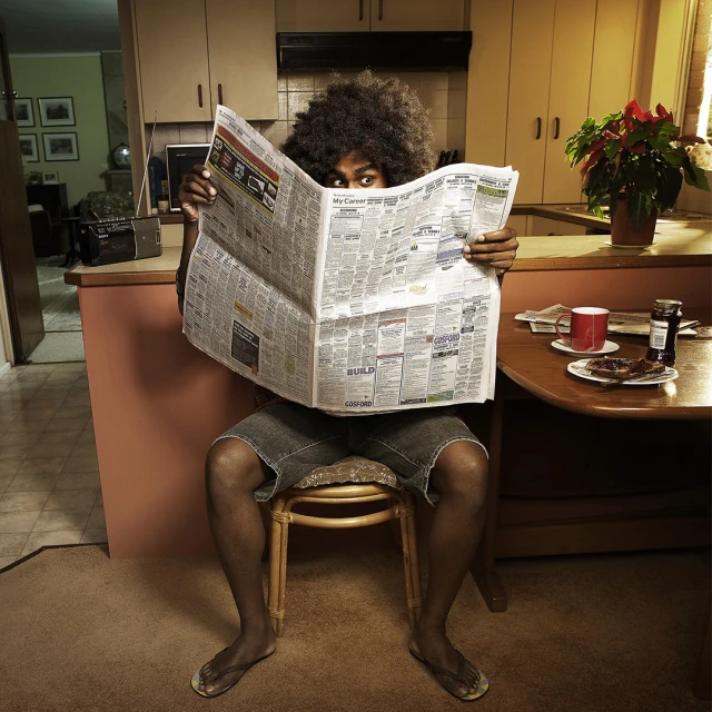 Adult male sitting on chair, looking over newspaper, home interior with breakfast on table and kitchen in the background.