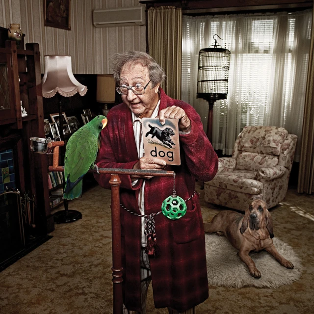 Elderly man in robe smiles holding flashcard with dog image; green parrot sits on perch with bloodhound dog in living room.