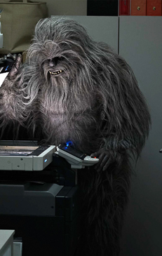 Furry, primate-like creature standing at a printer in a grey-walled office setting, illuminated by fluorescent lighting. 