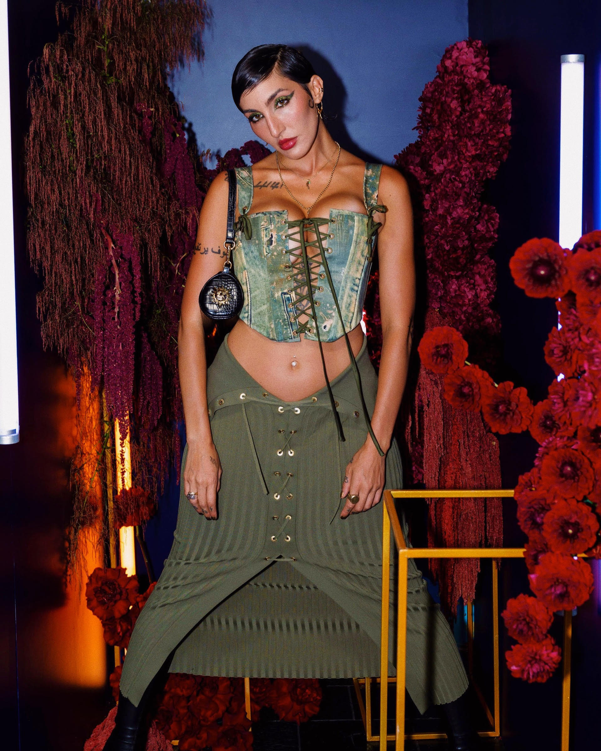 Woman in green corset and skirt poses in front of floral decorations and glowing lights, showcasing fashion and style.