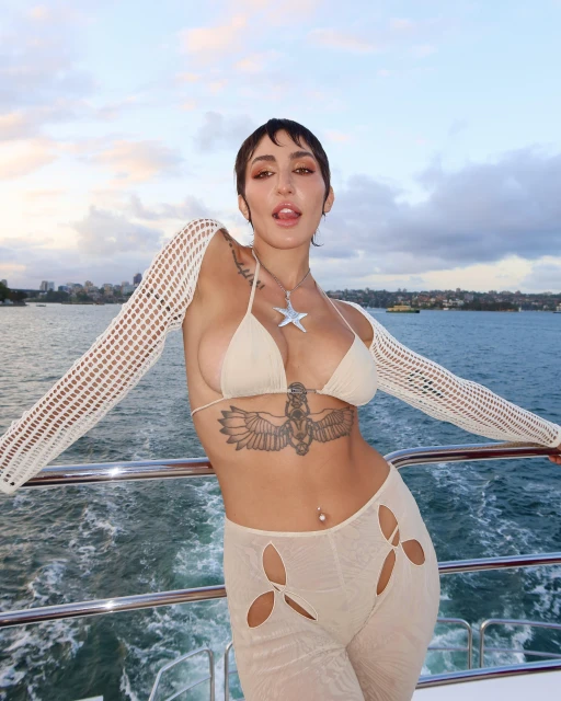 Adult woman poses on a boat wearing a bikini top, see-through pants, and long crochet sleeves with city buildings in the background.