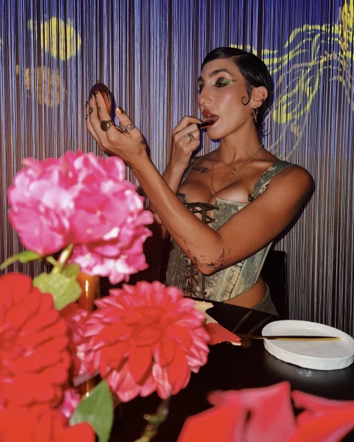 Young woman with short hair applies lipstick in an indoor setting with floral decorations and patterned curtains in the background.
