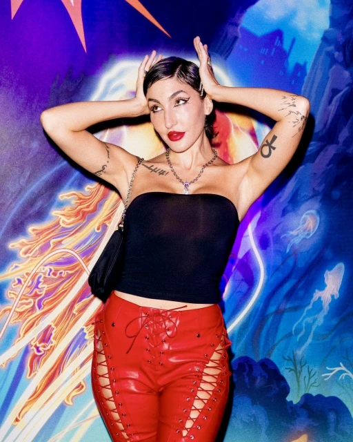 Woman with short dark hair poses in a black tube top and red leather lace-up pants against a painted backdrop of blue and purple.