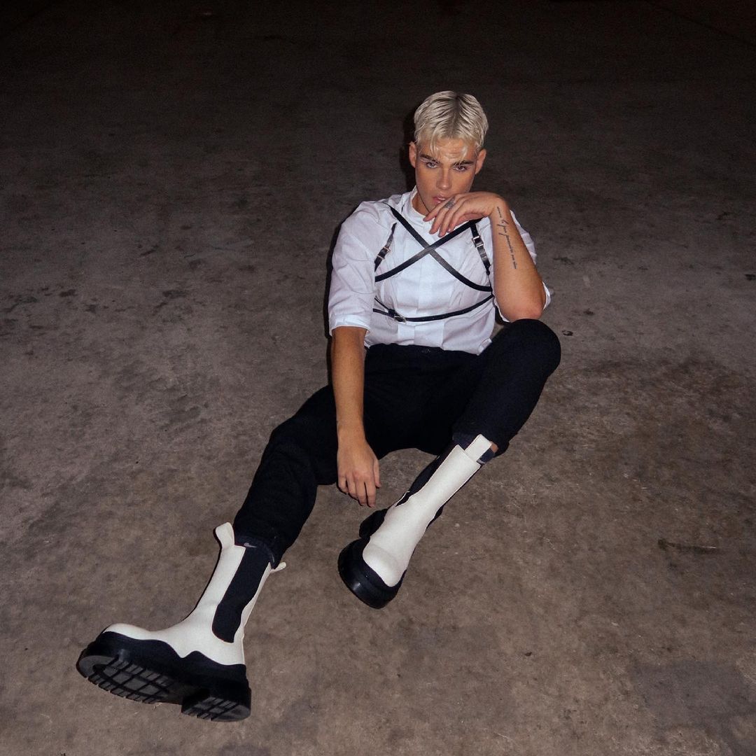 Young adult in a white shirt, black pants, and statement boots sitting on the ground in a dimly lit outdoor setting.
