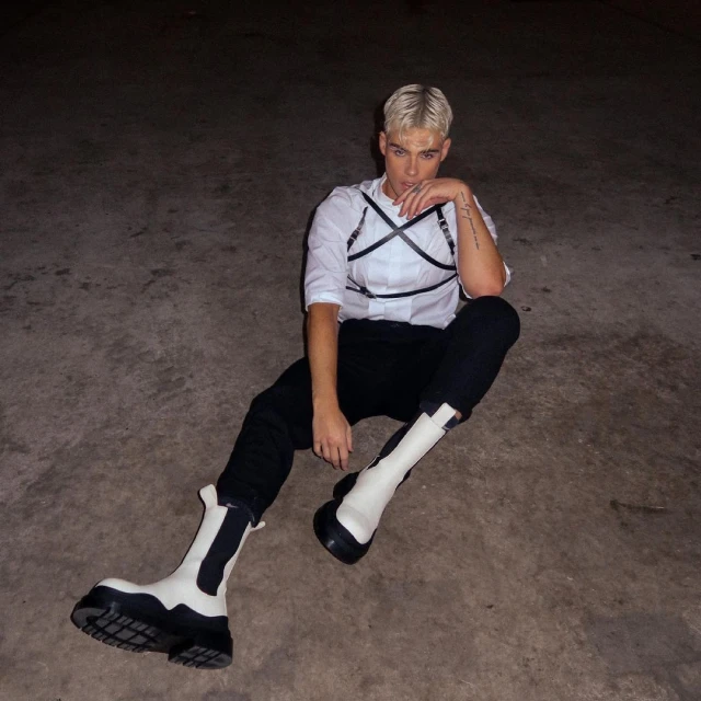 Young adult in a white shirt, black pants, and statement boots sitting on the ground in a dimly lit outdoor setting.