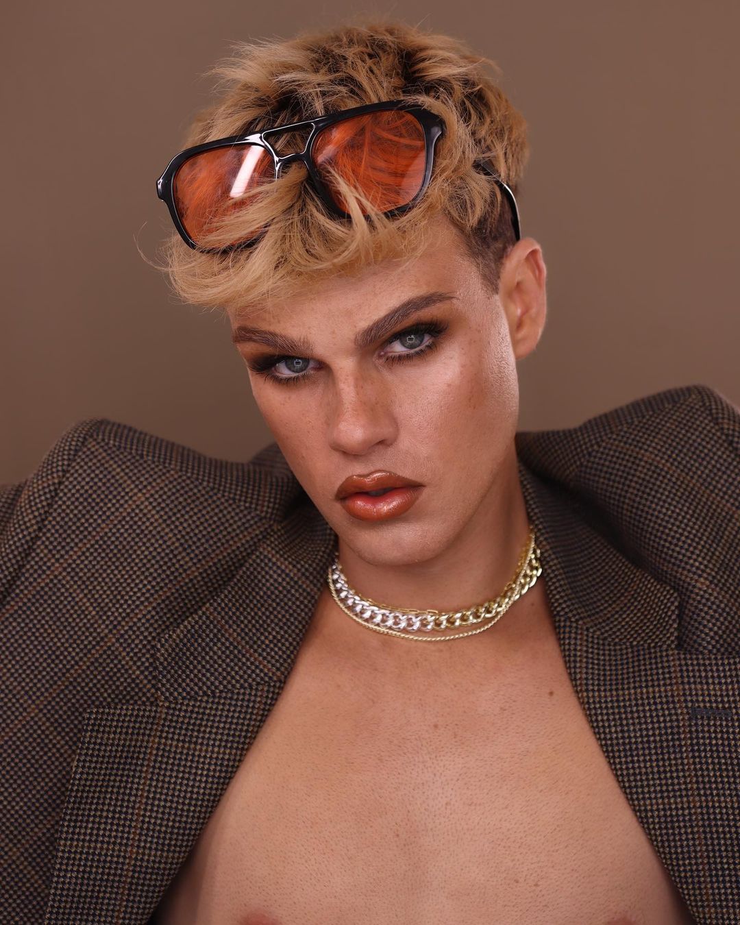 Glamorous portrait of a person with blonde hair wearing sunglasses and a blazer, showcasing bold makeup and chain necklaces.