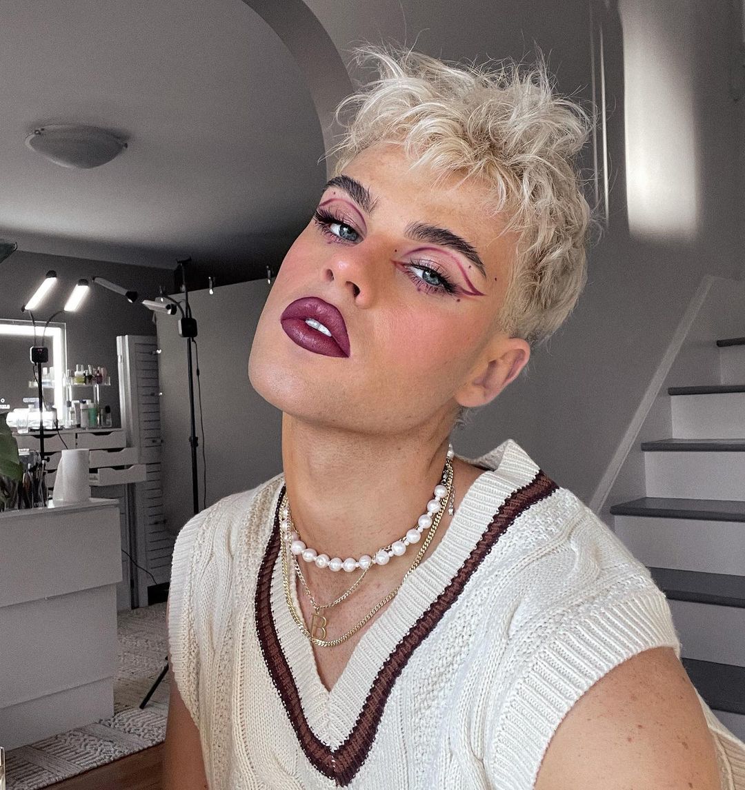 Person with short blond hair, wearing a white knit sweater vest and layered necklaces, poses in an indoor setting with makeup lights.
