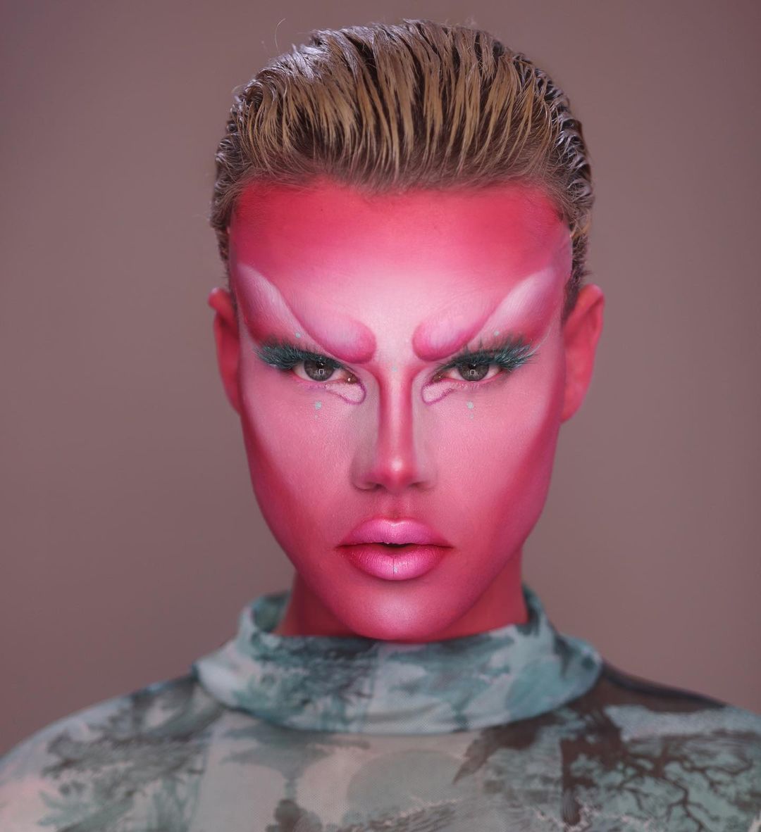 Close-up portrait of person with pink facial makeup, dramatic eyebrows, and green eyelashes. Wearing a teal patterned high-neck top.