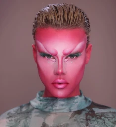 Close-up portrait of a person with vibrant pink makeup and slicked-back blonde hair wearing a patterned top against a neutral background.