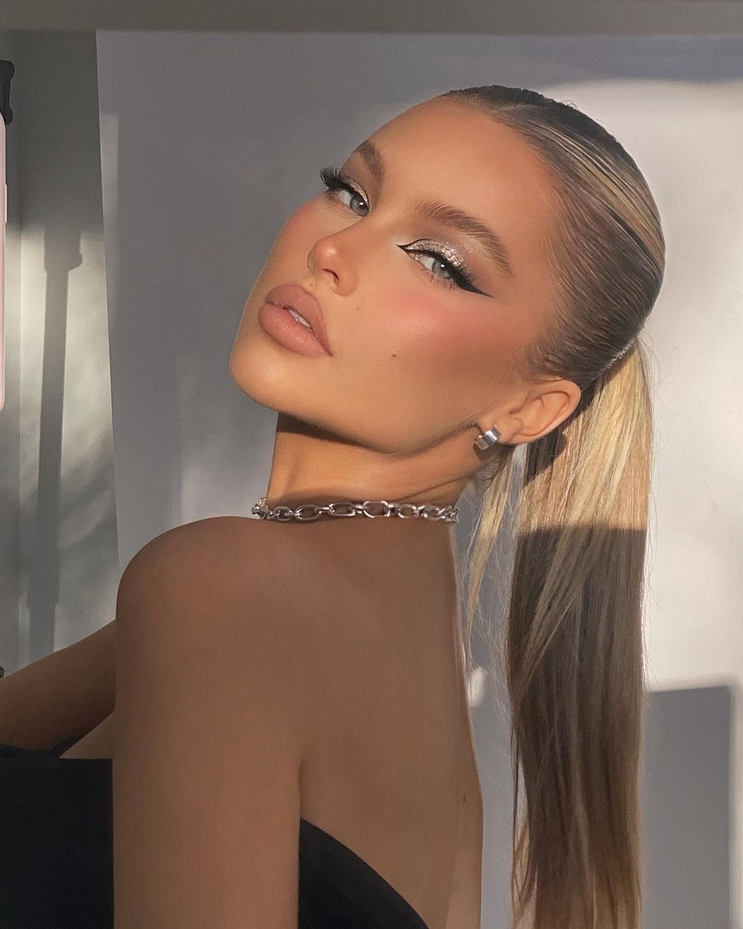 Close-up of an adult female with blond hair in a ponytail, wearing a chain necklace and dramatic makeup, posing in an indoor setting.