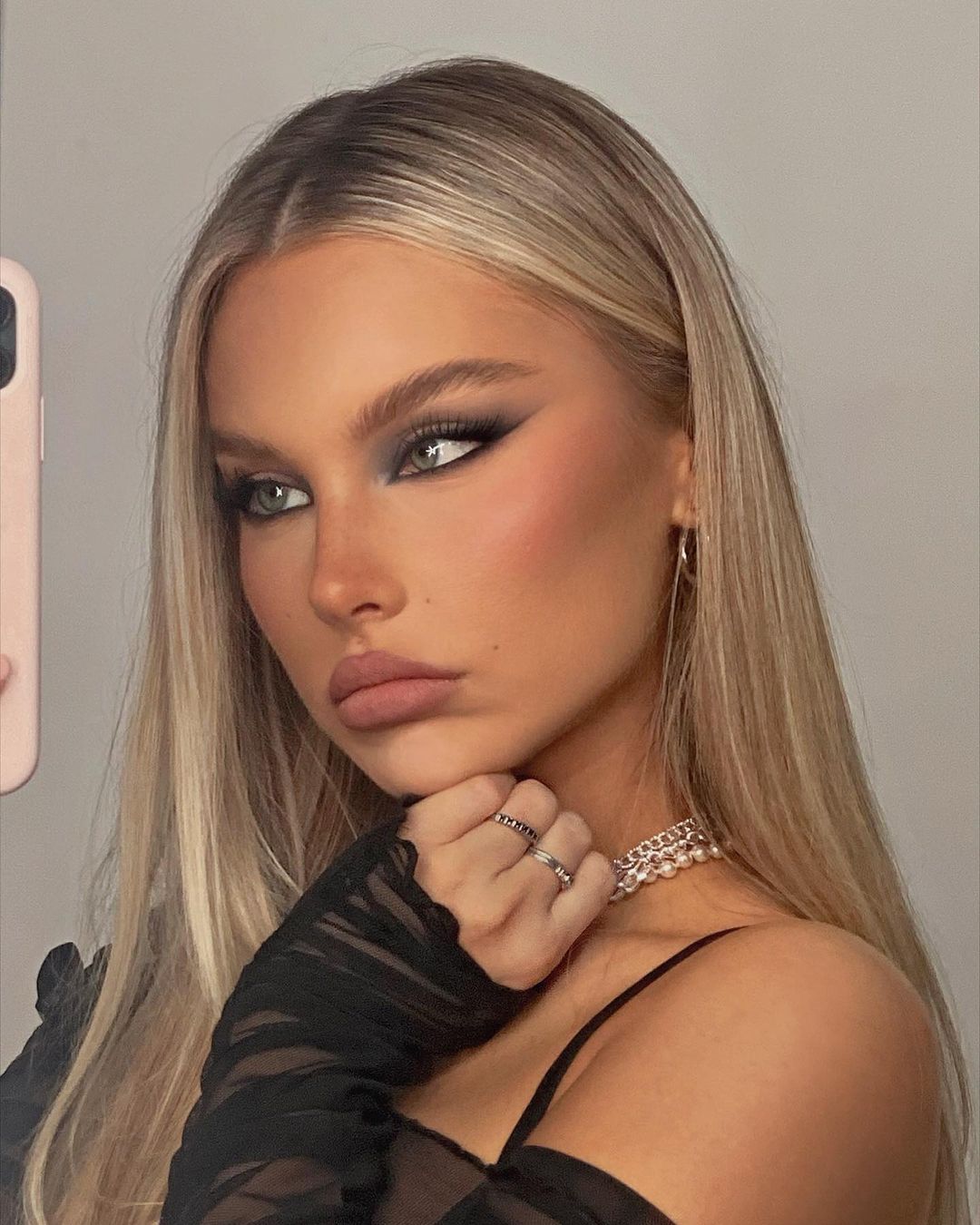 A young woman with blonde hair and gray smoky eye makeup poses indoors with a delicate sheer top and layered chain necklaces.