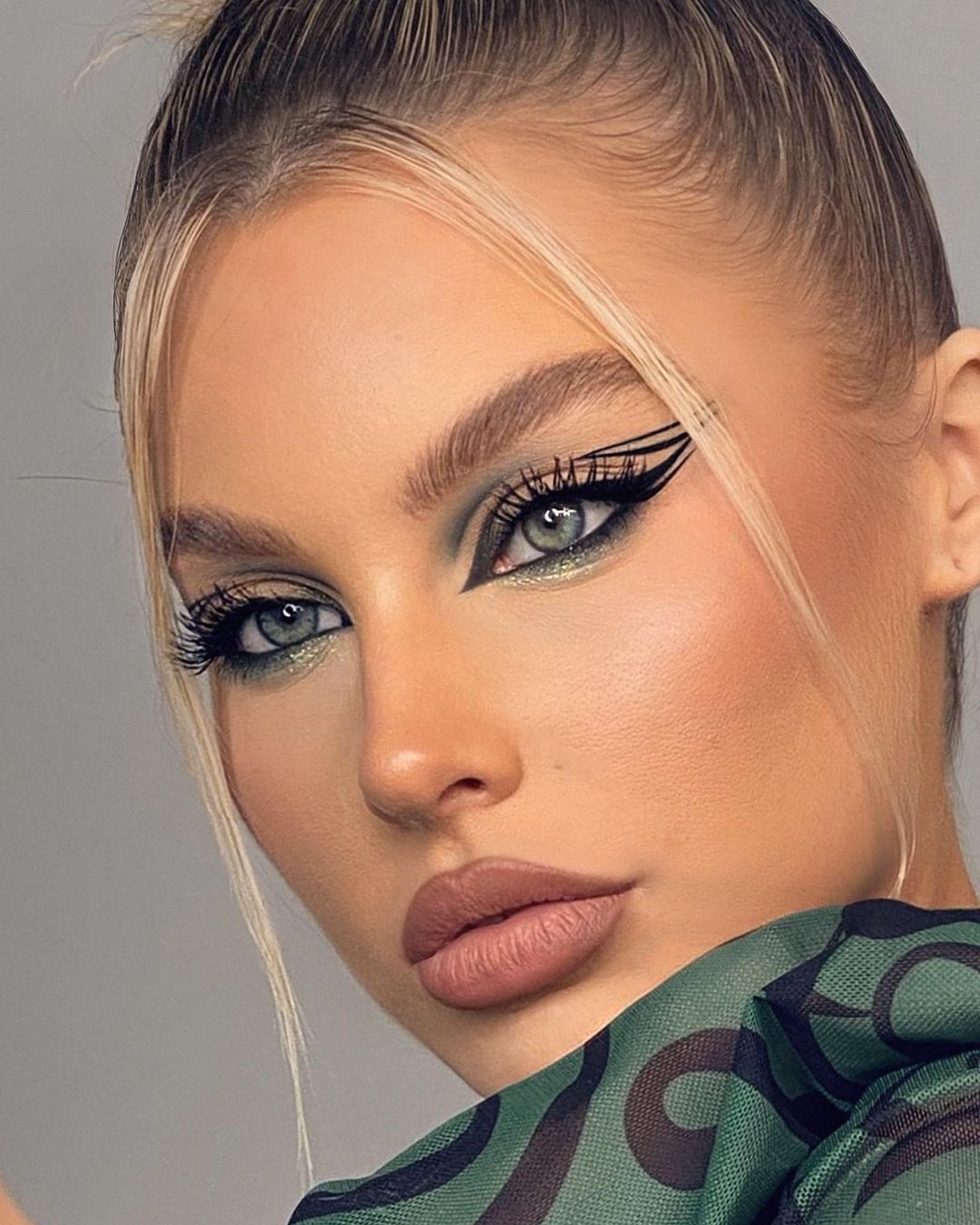 Close-up of a woman with dramatic graphic black winged eyeliner and green eyeshadow, wearing a green camouflage pattern mesh top.