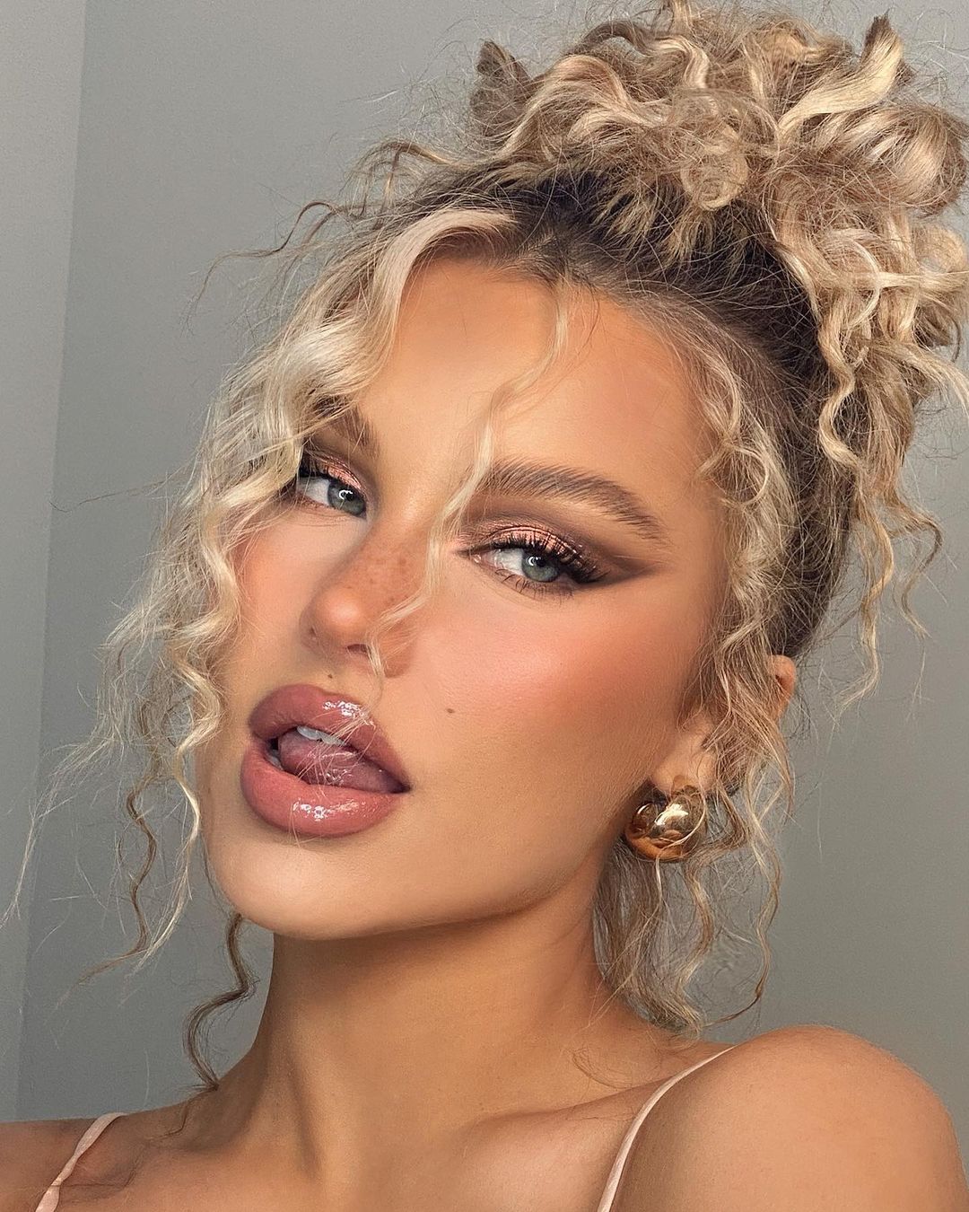 Close-up portrait of a blonde woman with curly hair styled in a messy bun, wearing gold earrings and makeup with a grey background.
