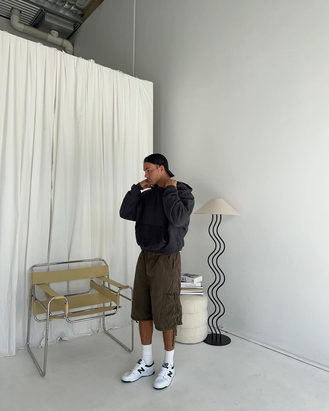 An adult male stands in a studio wearing a hoodie, shorts, socks, and sneakers, next to a lamp, chair, and stacked books against a white background.