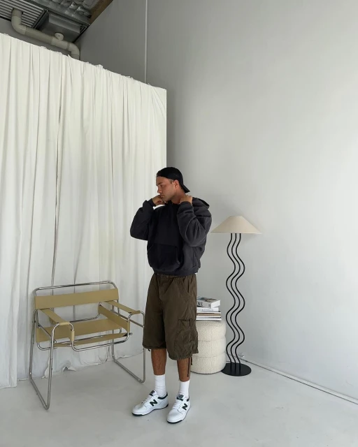 An adult male stands in a studio wearing a hoodie, shorts, socks, and sneakers, next to a lamp, chair, and stacked books against a white background.