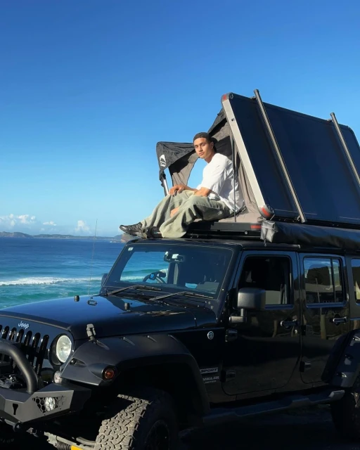 Adult male sits atop a black SUV with a pop-up camper, ocean backdrop, clear blue sky, sunny outdoor environment, and rugged terrain.