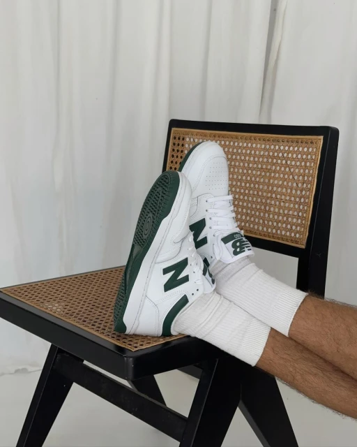 Person wearing white New Balance sneakers with green accents, sitting on a black and tan wicker chair in front of a white curtain.