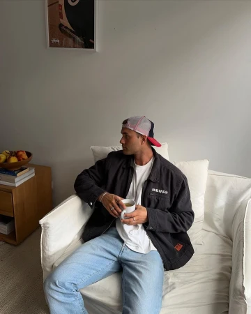 Man wearing a dark jacket and baseball cap seated on a white sofa holding a white mug in a light interior.