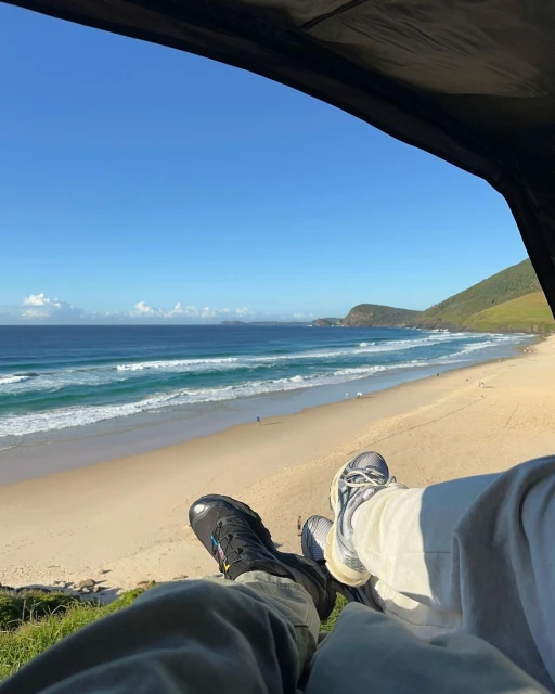 View from inside a tent of a sandy beach and ocean with waves on a sunny day, seen with two pairs of feet in the foreground.