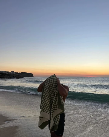 Person dries with a checkered towel at sunrise on a beach with ocean waves and a building on a cliff visible in the background.