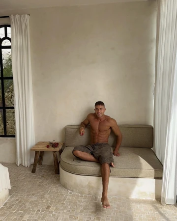 Shirtless man sitting on a beige upholstered bench in a bright minimalist room with white sheer curtains and stone tiled floor