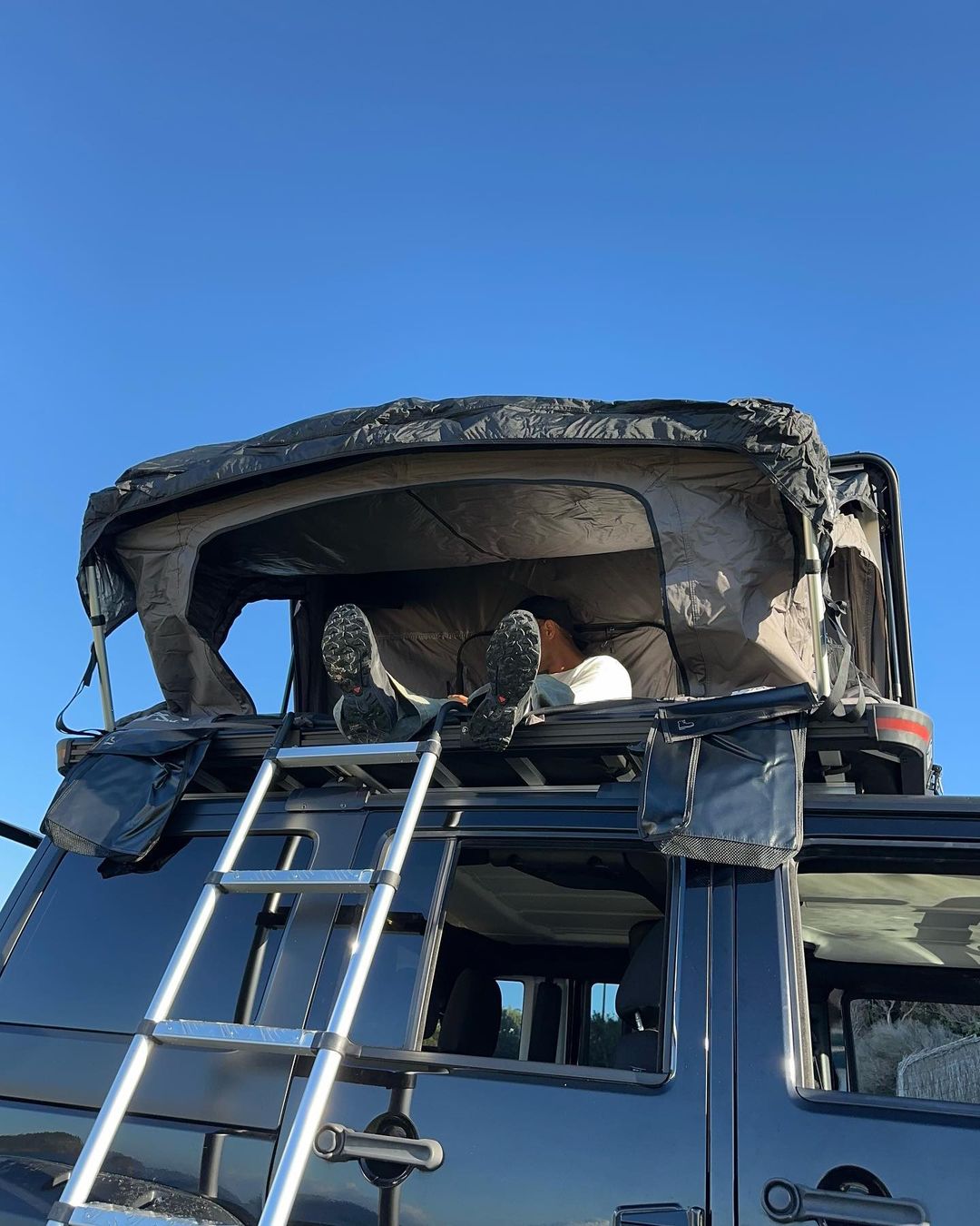Person relaxing on a rooftop tent on a car, with a ladder propped against the vehicle under a clear blue sky in a daylight scene.