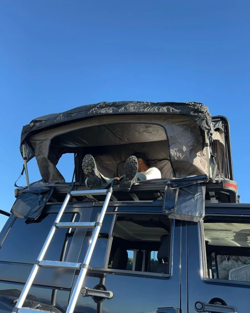 Person relaxing on a rooftop tent on a car, with a ladder propped against the vehicle under a clear blue sky in a daylight scene.