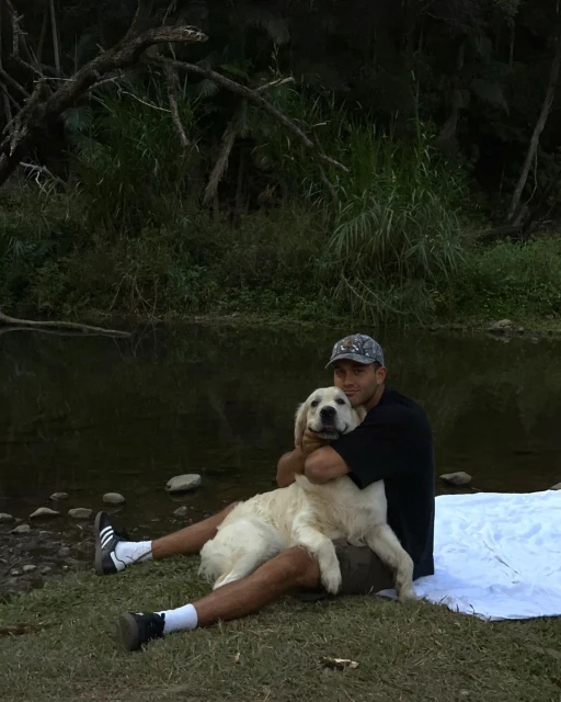 Man hugging a Golden Retriever dog outdoors near a river, sitting on the grass with a white blanket. The dog is light-furred and relaxed.