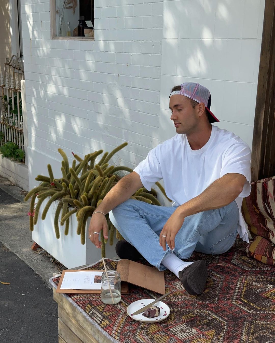 An adult male sits at an outdoor cafe table with food and drink in the daylight, wearing casual clothing and a baseball cap in a candid shot.
