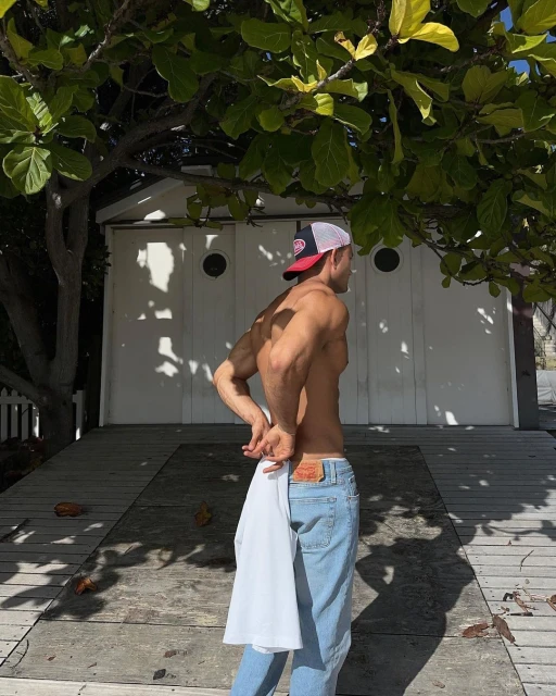 Fit adult male stands shirtless, wearing jeans and a cap, holding a white t-shirt in an outdoor setting with trees and a small white building.