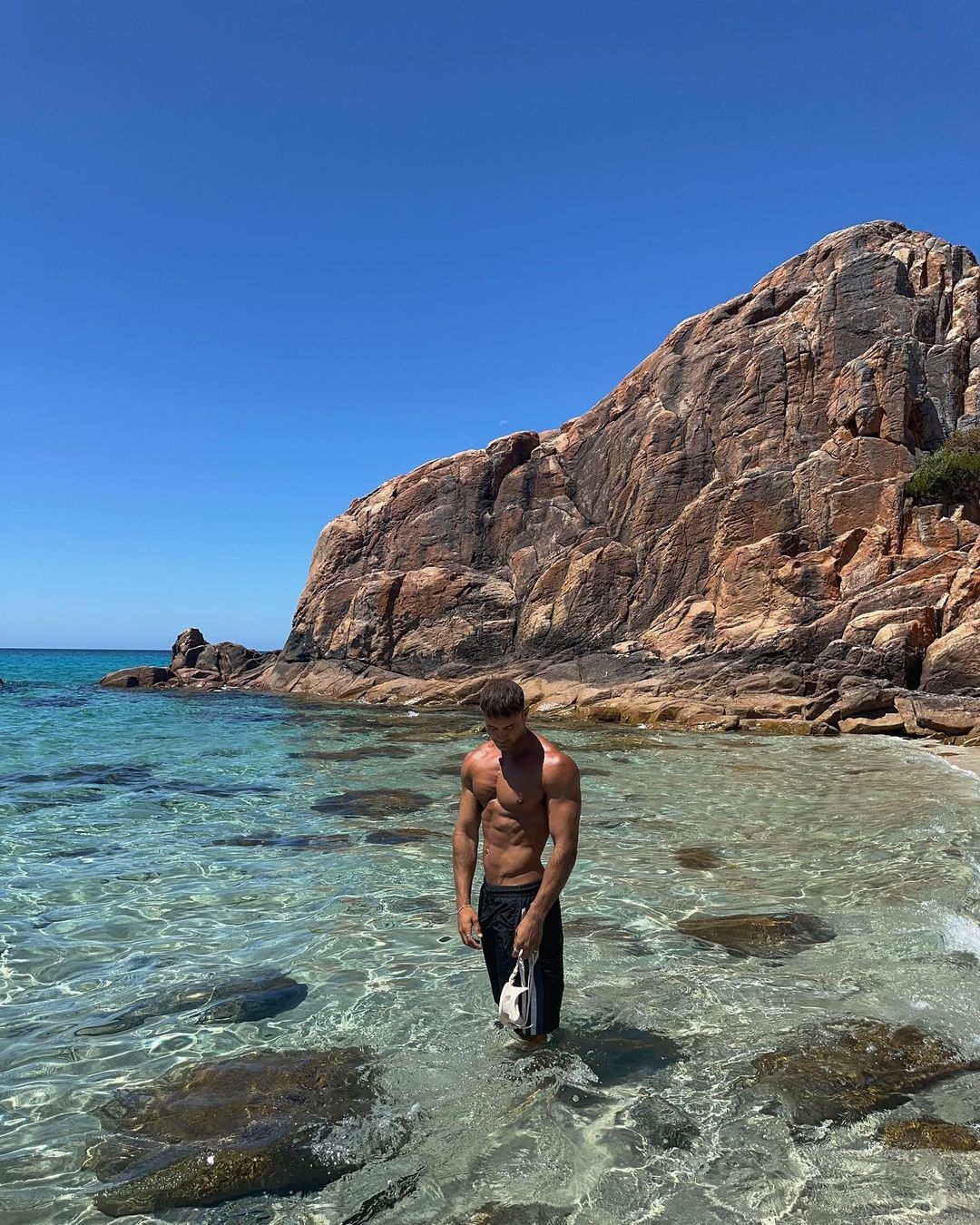 Adult male stands in clear ocean water on a sunny day with a rocky cliff backdrop. He is holding a bag.