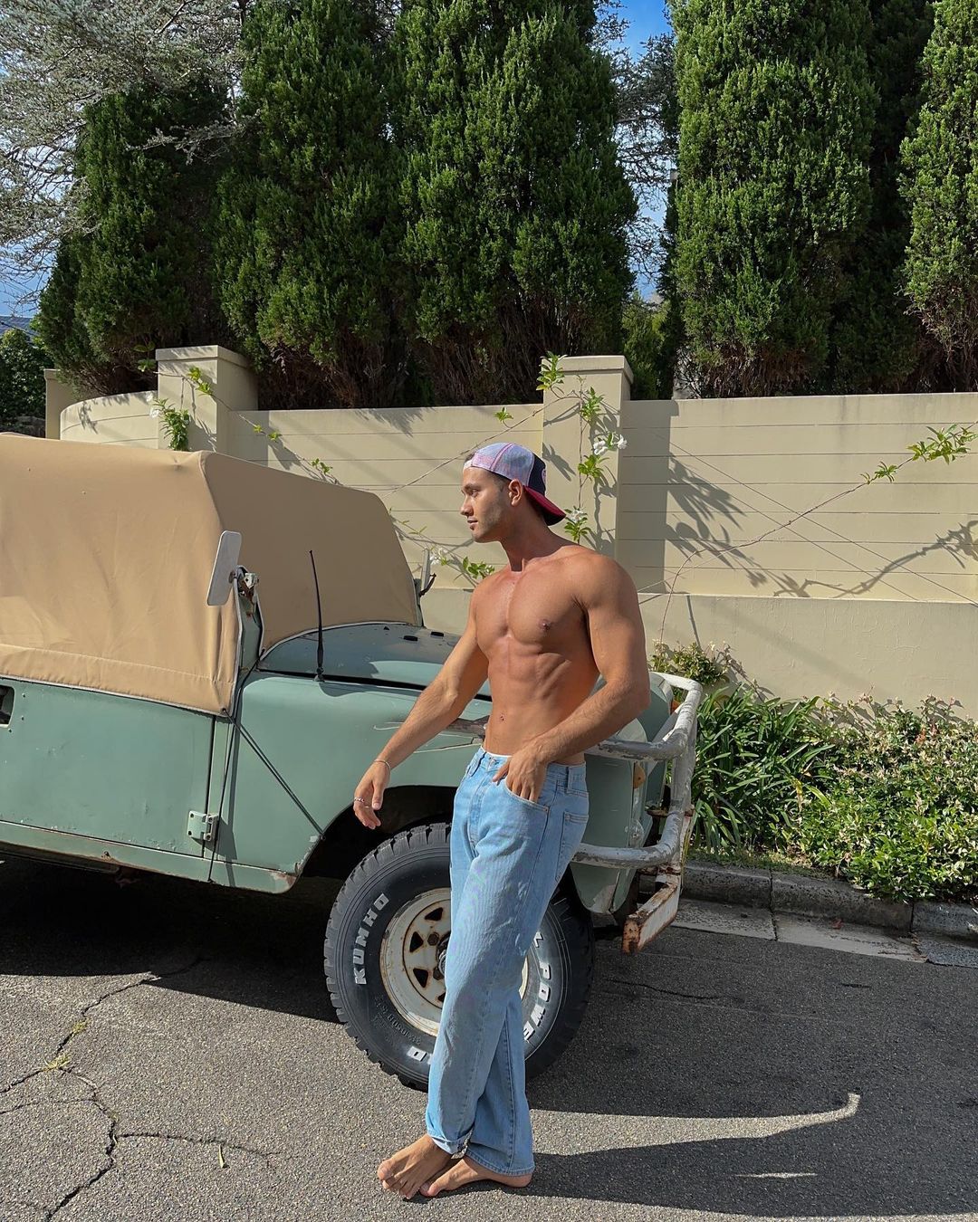 Shirtless man in jeans and cap poses next to an olive green jeep on an asphalt road with trees and a tan wall in the background.