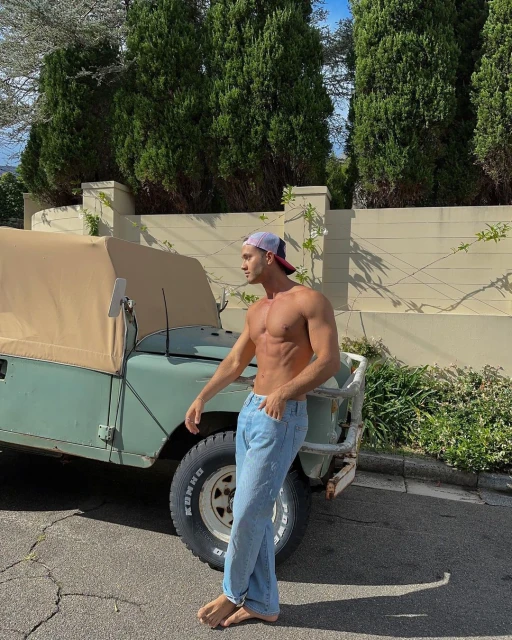 Shirtless man in jeans and cap poses next to an olive green jeep on an asphalt road with trees and a tan wall in the background.
