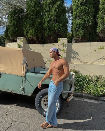 Shirtless man in jeans and cap poses next to an olive green jeep on an asphalt road with trees and a tan wall in the background.