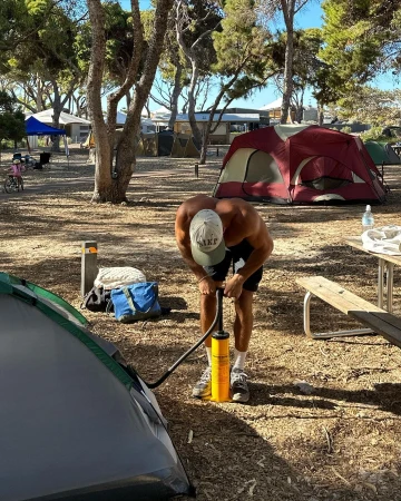 Adult male pumping a tent at a campsite with other tents and trees in the background under a blue sky on a sunny day.