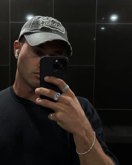 Selfie of a young adult man wearing a baseball cap and rings, holding a phone in an indoor setting with dark tiled background.